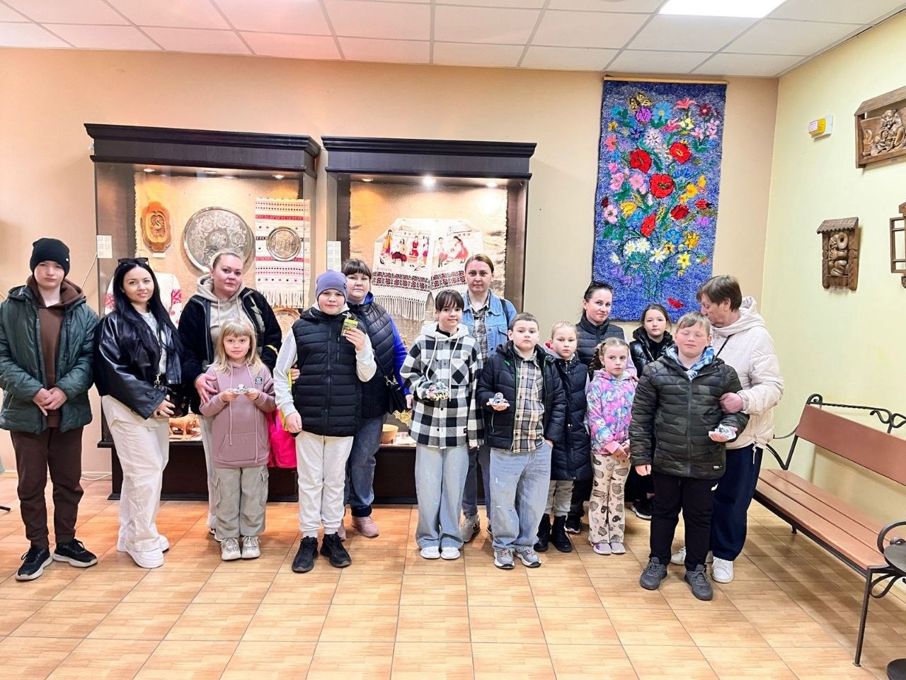 Група дорослих та дітей у верхньому одязі стоїть у приміщенні музею чи виставкової зали. На тлі видно скляні вітрини з експонатами народної творчості, вишитий рушник, а також яскравий гобелен із квітами на стіні.