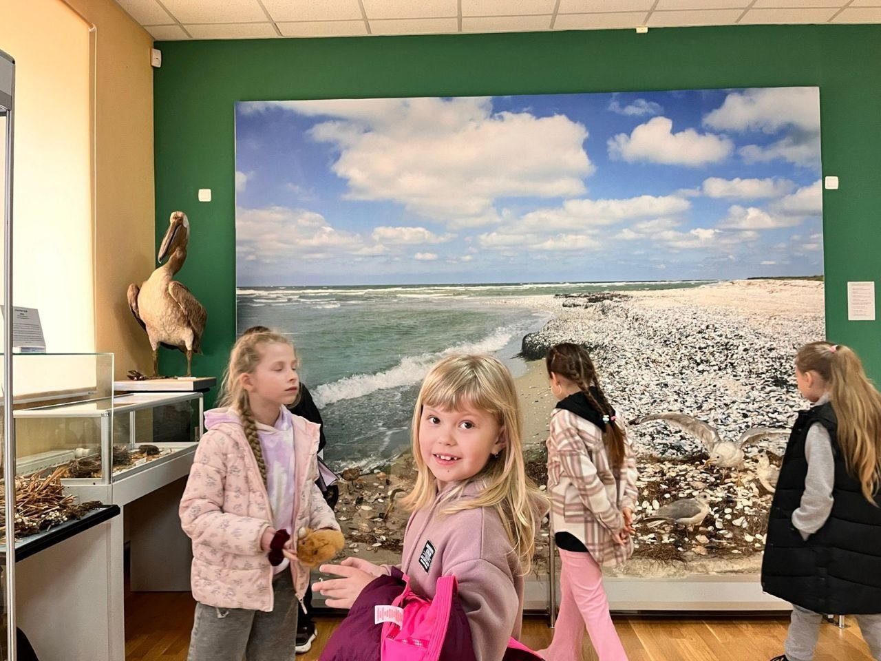 Діти в музеї на тлі великої панорамної фотографії морського узбережжя та неба з хмарами. Ліворуч видно скляну вітрину та опудало великого птаха.