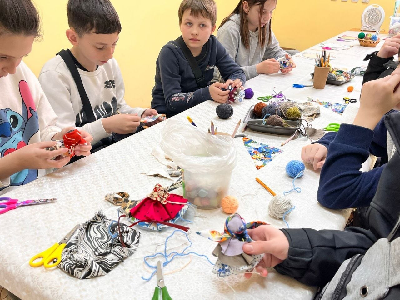 Діти сидять за довгим столом і виготовляють ляльки з кольорових тканин та ниток під час творчого заняття. На столі розкладені клубки пряжі, ножиці та клаптики матеріалу.