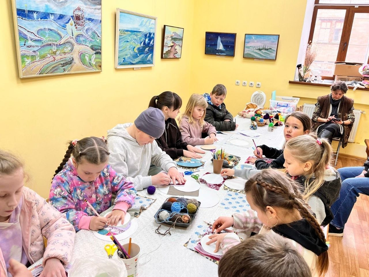 Група дітей сидить за довгим столом у світлій кімнаті та займається творчістю під час майстер-класу.