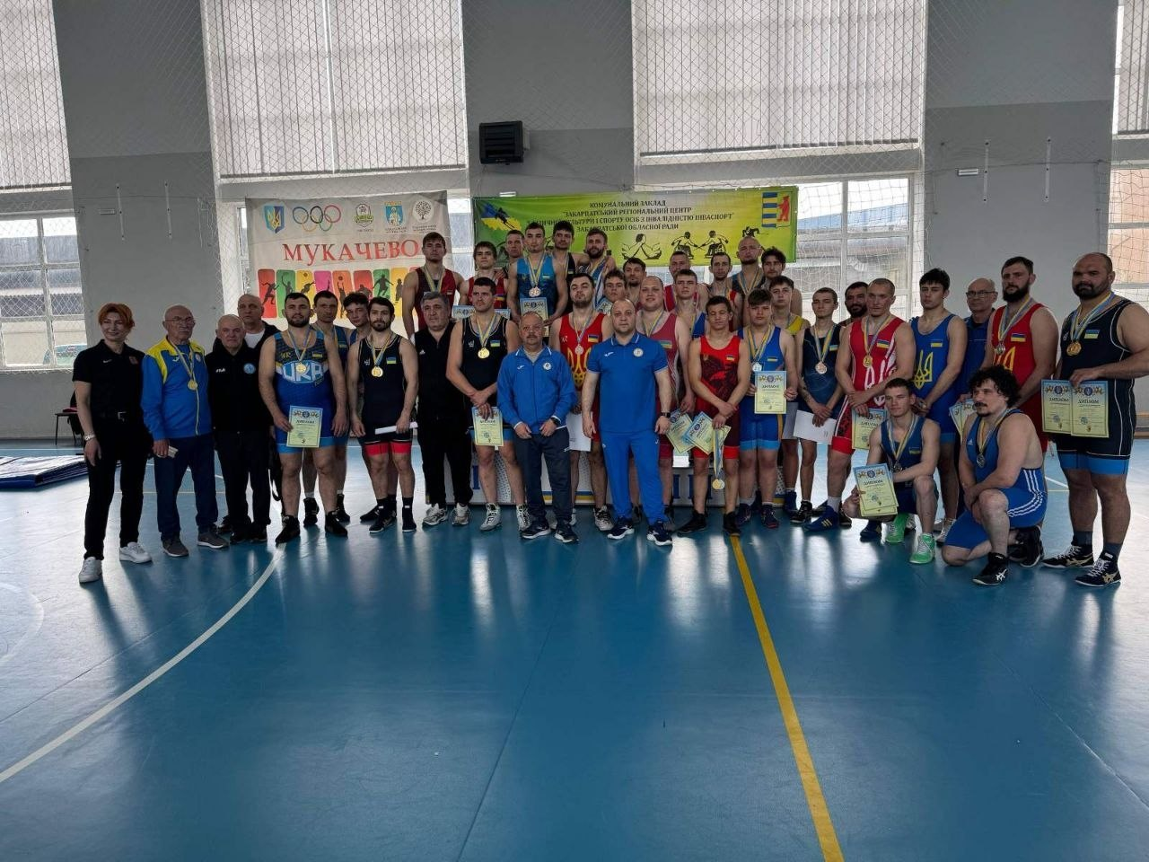 Велика група спортсменів та тренерів у спортивній формі стоїть на майданчику у спортивному залі. Багато учасників тримають у руках грамоти та мають медалі на шиї. На задньому плані видно великі вікна, а також банери з написом «Мукачево» та державною символікою.