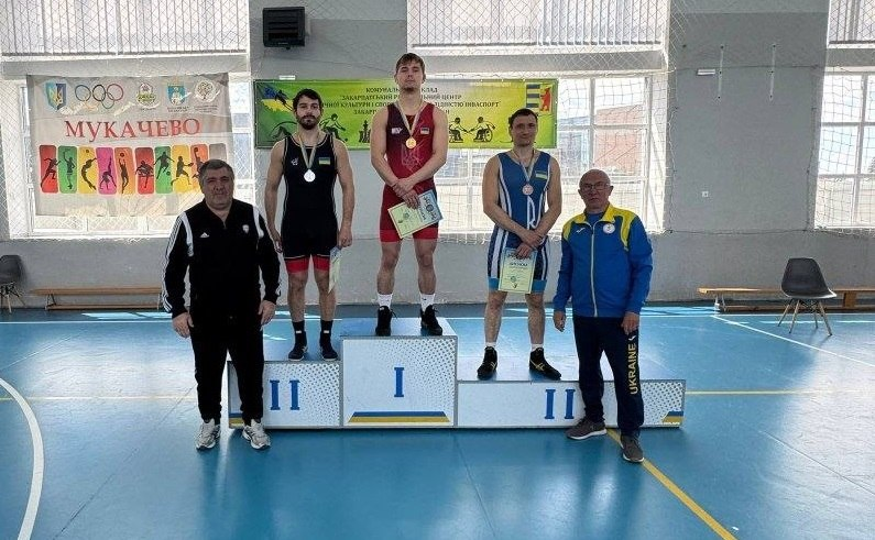 Троє борців у чорному, червоному та синьому трико стоять на п'єдесталі з медалями та грамотами. Поруч із ними стоять двоє чоловіків у спортивних костюмах у залі з банером «Мукачево».