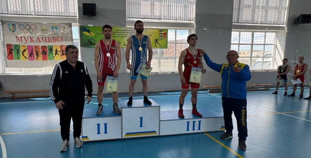 Троє борців у трико стоять на п'єдесталі з медалями. На першому місці спортсмен у синьо-жовтому трико. Другий та третій призери мають червоні трико. Поруч стоять двоє чоловіків на тлі банера «Мукачево».
