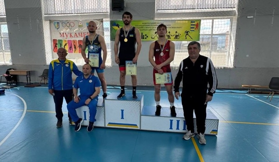 Троє борців у синьому, чорному та червоному трико стоять на п'єдесталі з медалями та грамотами в руках. Поруч один чоловік сидить, а двоє стоять у спортивних костюмах на тлі банера «Мукачево».