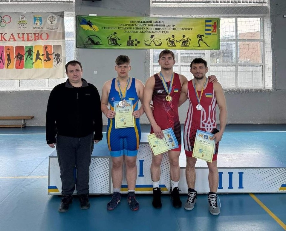 Троє борців у синьому, темно-червоному та червоному трико з білим тризубом стоять із медалями та дипломами в руках. Ліворуч від них стоїть чоловік у чорній кофті. Усі вони перебувають у спортивній залі на тлі п'єдесталу та інформаційних банерів.