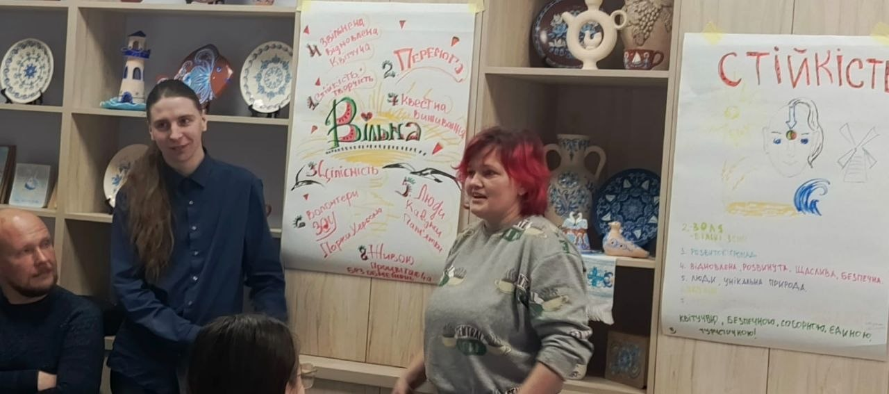 Жінка з яскраво-червоним волоссям виступає на тлі стелажів із керамічним посудом та декоративними тарілками. На меблях закріплені два великі паперові плакати з малюнками та написами «Вільна» та «Стійкість». Поруч стоять та сидять інші учасники заходу.