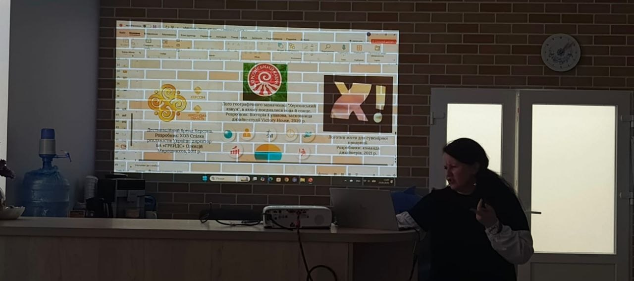 Жінка проводить презентацію в приміщенні за допомогою ноутбука та проєктора. На цегляну стіну проєктується слайд із текстом та різними логотипами, що стосуються брендингу міста Херсон. Ліворуч на столі стоїть великий бутель із водою.