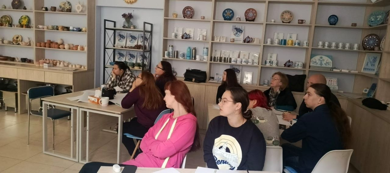 Група людей сидить за столами під час лекції або майстер-класу в приміщенні. На задньому плані розташовані великі дерев'яні стелажі з виставленими керамічними тарілками, чашками та іншими сувенірами.