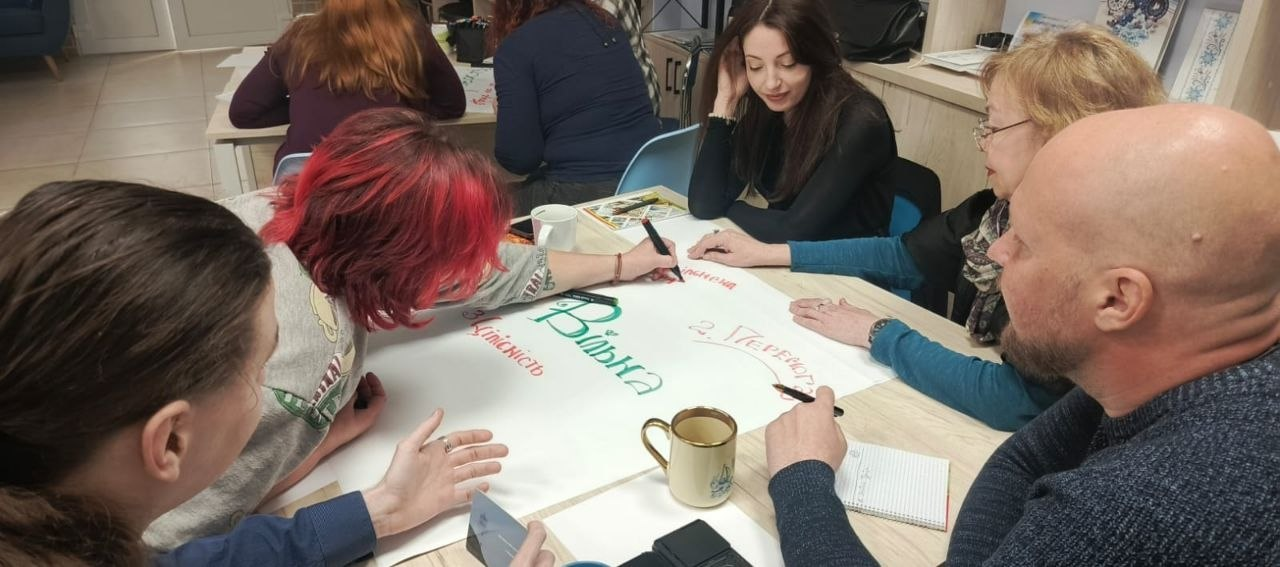 Група людей сидить за столом і спільно працює над створенням плаката на великому аркуші паперу. Жінка з яскравим червоним волоссям пише зелене слово «Вільна». Інші учасники спостерігають та роблять нотатки.