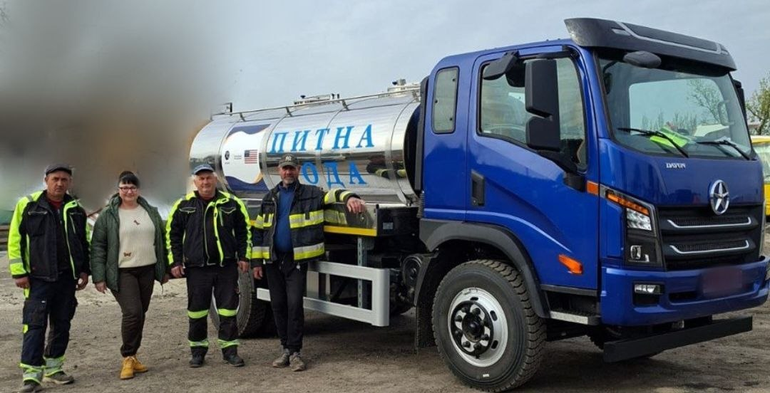 Чотири людини стоять біля великої синьої вантажівки з металевою цистерною, на якій нанесено великий блакитний напис «ПИТНА ВОДА». Три чоловіки та жінка одягнені в повсякденні та робочі куртки. На сріблястій цистерні також видно прапор США та емблему гуманітарної допомоги.