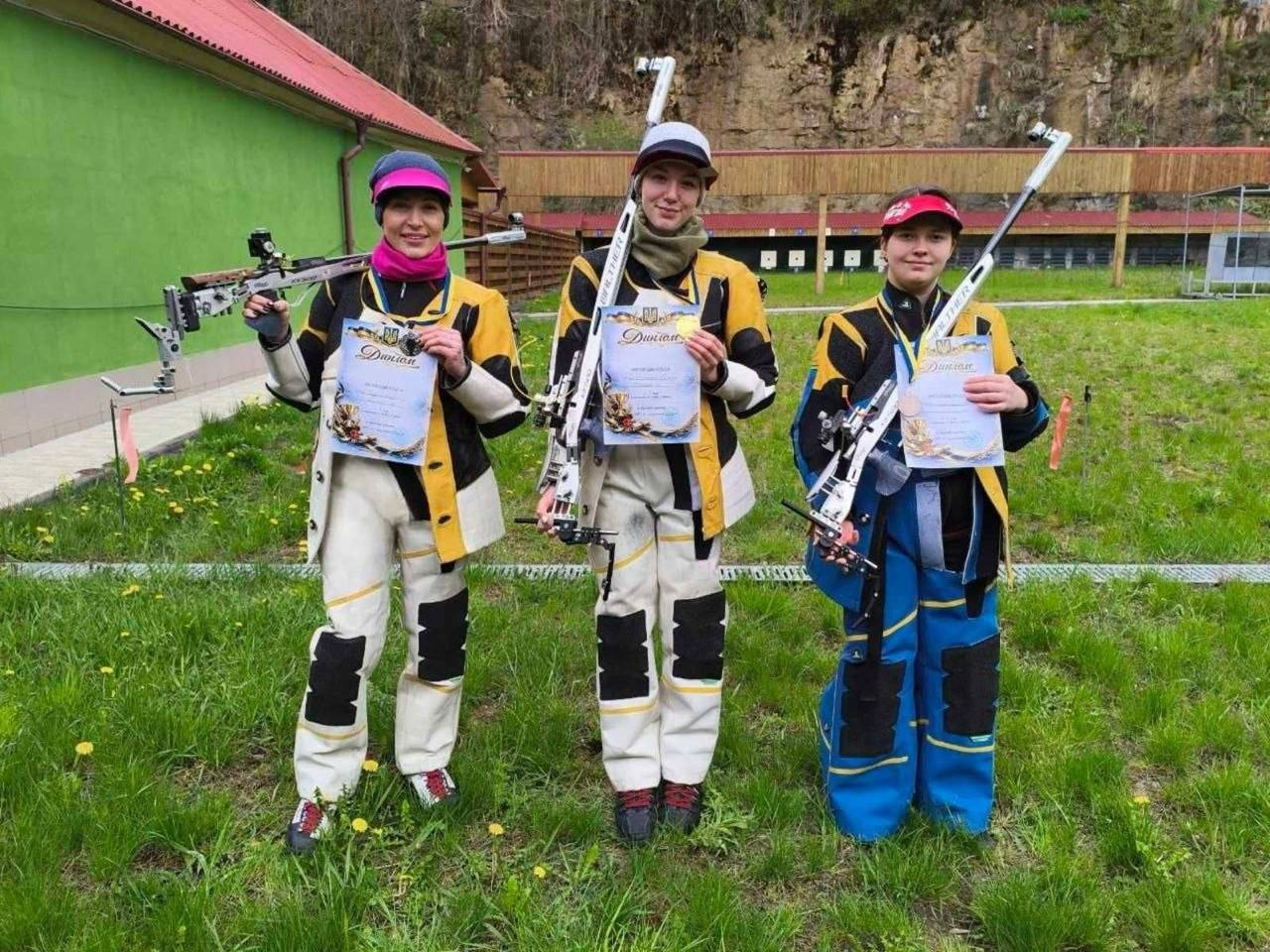 Три дівчини у спеціальних спортивних костюмах для кульової стрільби стоять на траві просто неба. Кожна з них тримає в руках спортивну гвинтівку та паперовий диплом. На задньому плані видно зелену будівлю та обладнаний стрілецький тир.