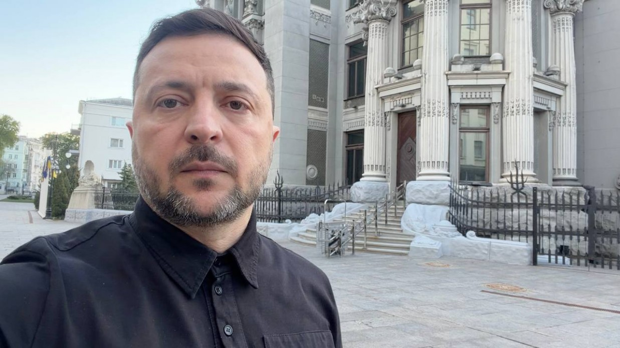 Чоловік у чорній сорочці робить селфі на вулиці на тлі величної сірої будівлі з масивними колонами та сходами.