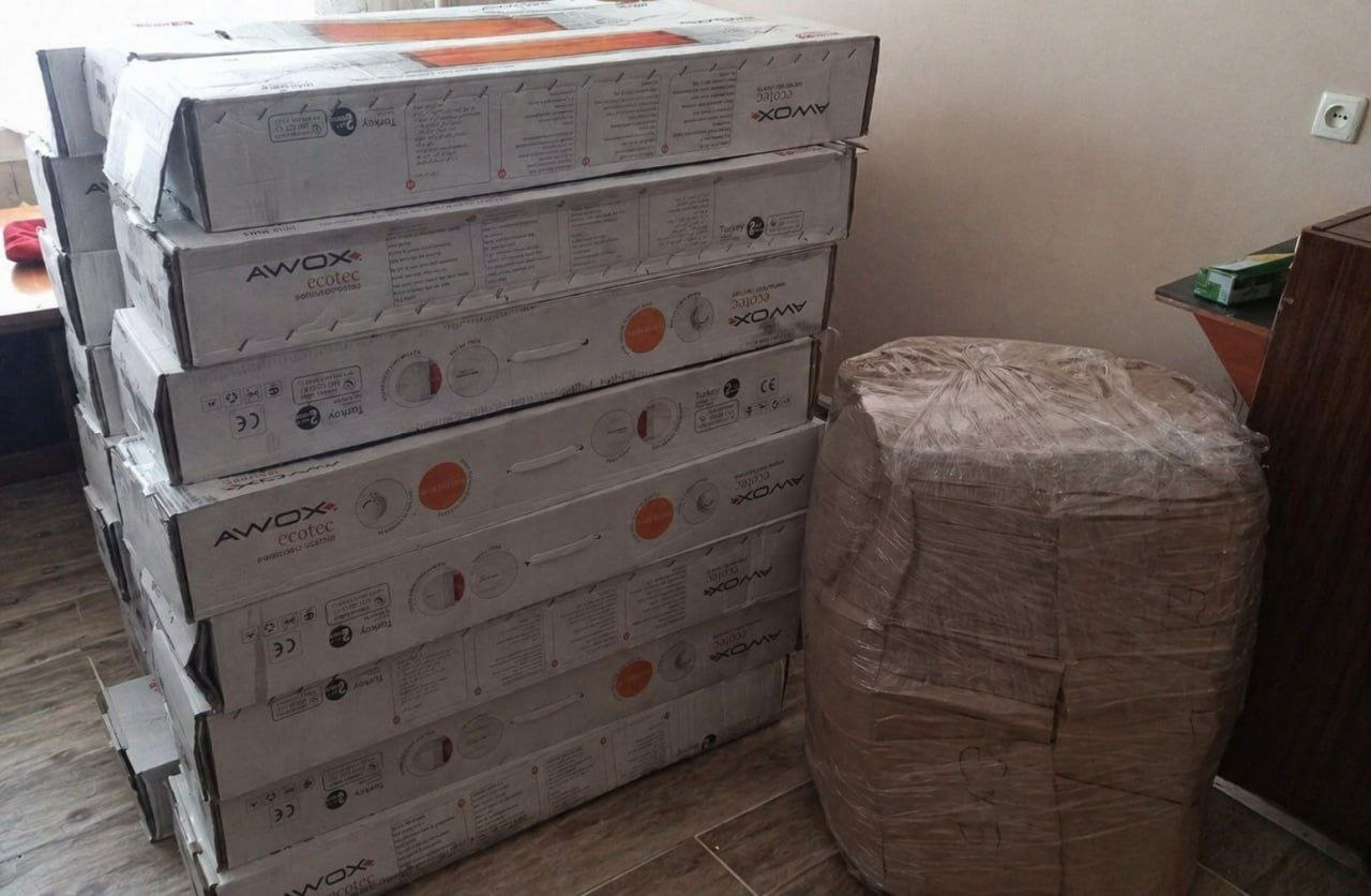 Стос довгих картонних коробок з написом «AWOX ecotec», складених одна на одну біля стіни в кімнаті. Поруч стоїть великий об'єкт циліндричної форми, щільно обмотаний пакувальною плівкою.