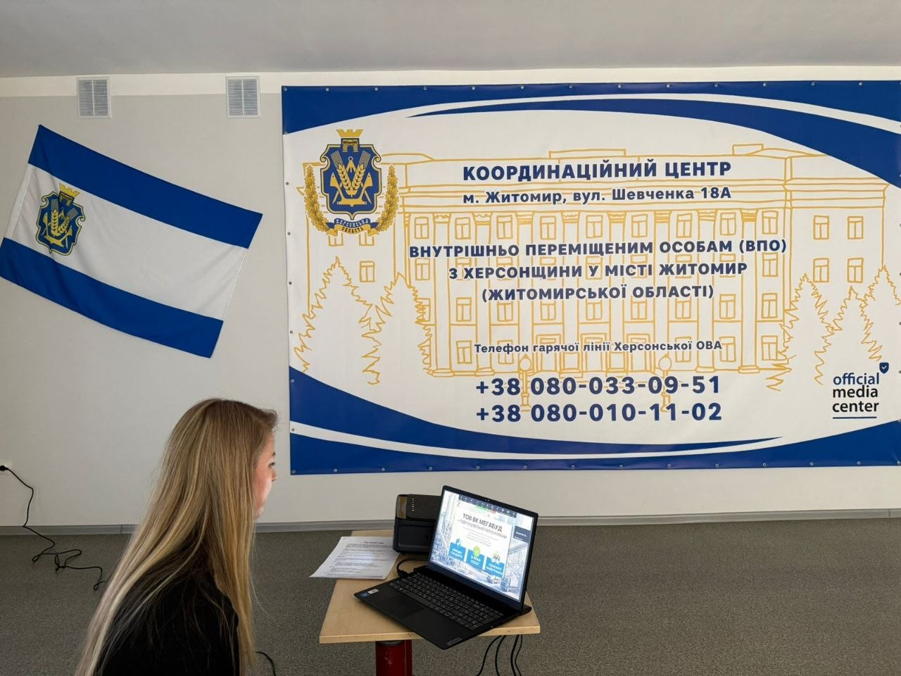 Дівчина за ноутбуком у волонтерському штабі на фоні банера з контактами гарячої лінії для допомоги внутрішньо переміщеним особам.