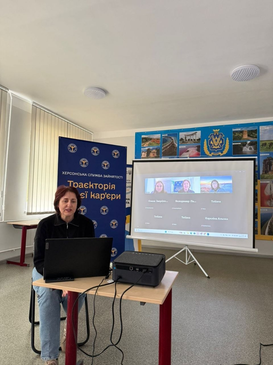 Жінка проводить онлайн-зустріч за ноутбуком. Поруч стоїть проєктор і банер служби зайнятості з написом «Траєкторія твоєї кар'єри», а на екрані видно учасників відеоконференції.