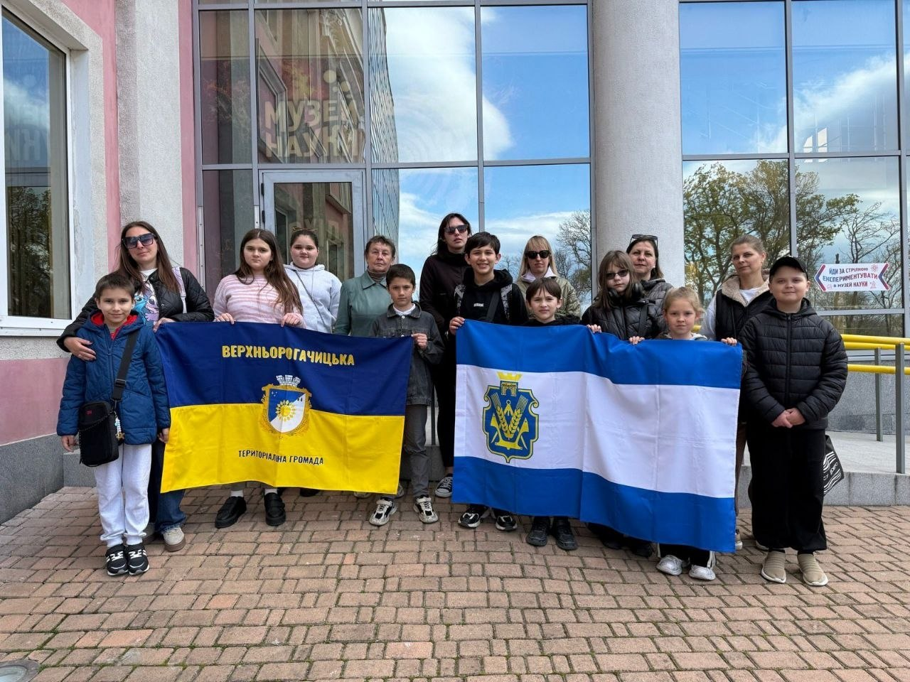 Група дітей та дорослих позує для спільного фото перед будівлею Музею науки. Вони тримають розгорнуті прапори Верхньорогачицької територіальної громади та міста Херсон.