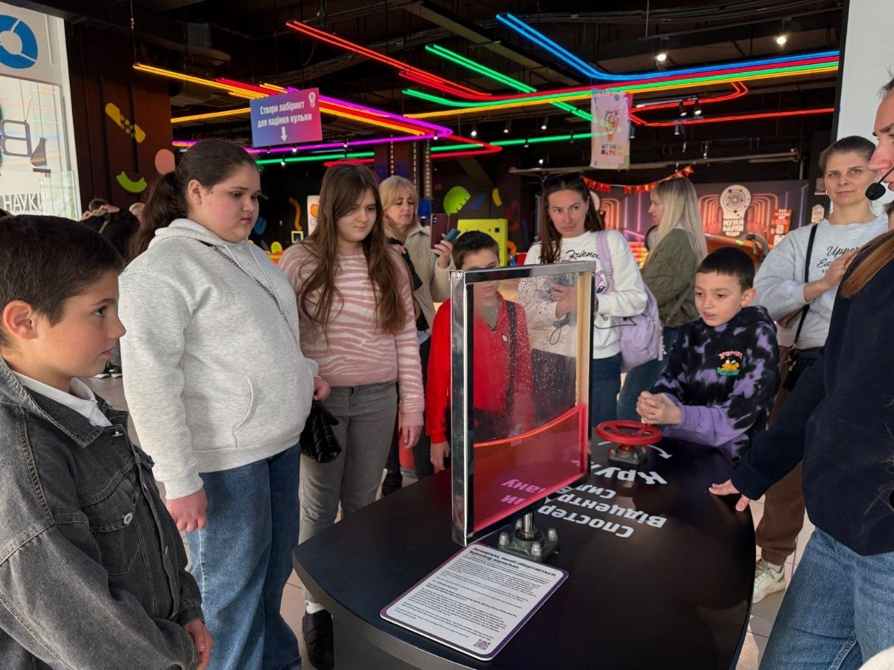 Група дітей та дорослих у музеї науки спостерігає за дією інтерактивного експоната з червоною рідиною. Один із хлопчиків крутить спеціальний вентиль, щоб привести механізм у дію, під наглядом екскурсовода.