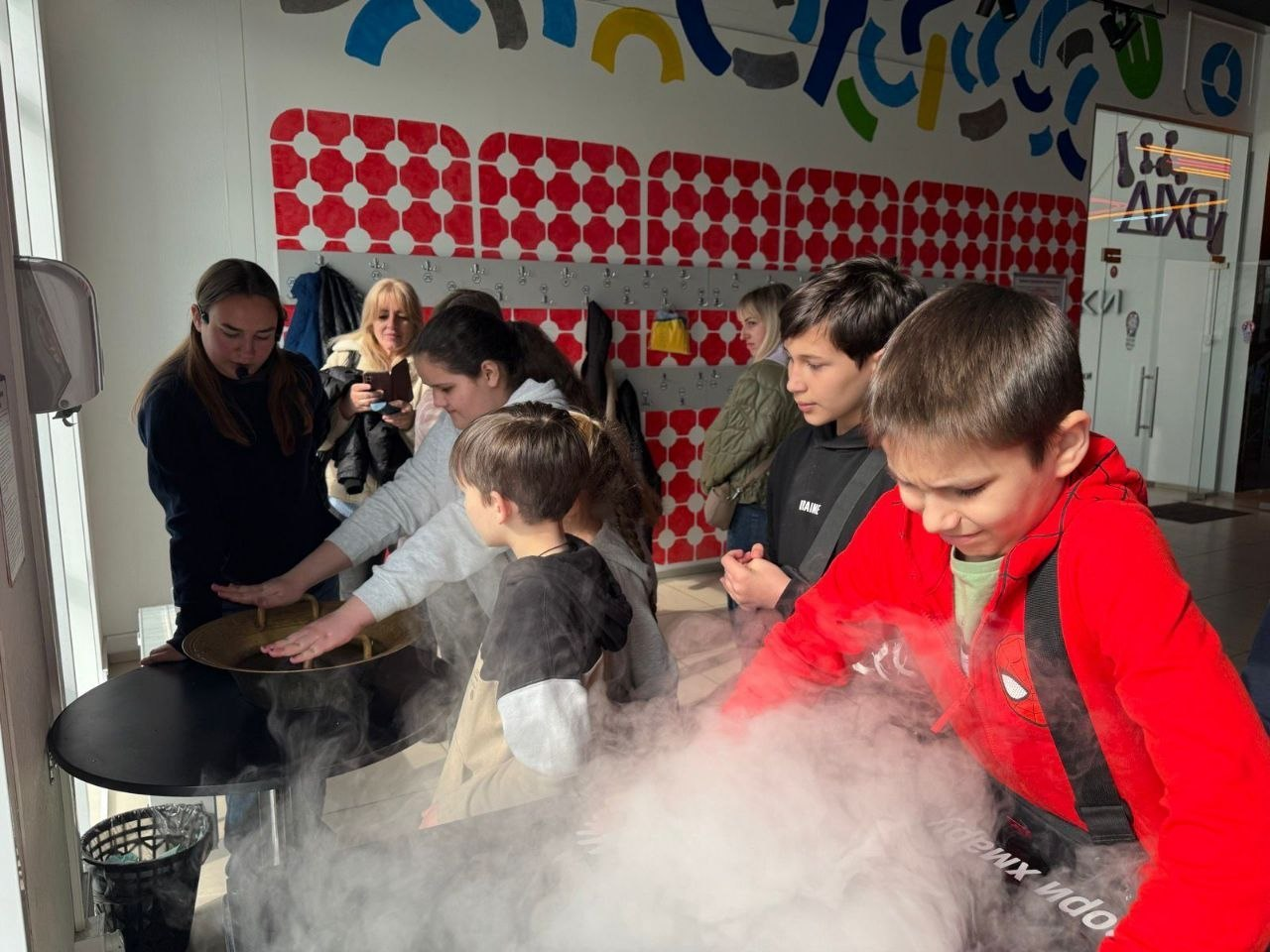 Група дітей та дорослих проводить науковий експеримент із густою білою парою, що розтікається по столу. Учасники з цікавістю спостерігають за процесом і торкаються пари руками в сучасному освітньому просторі.