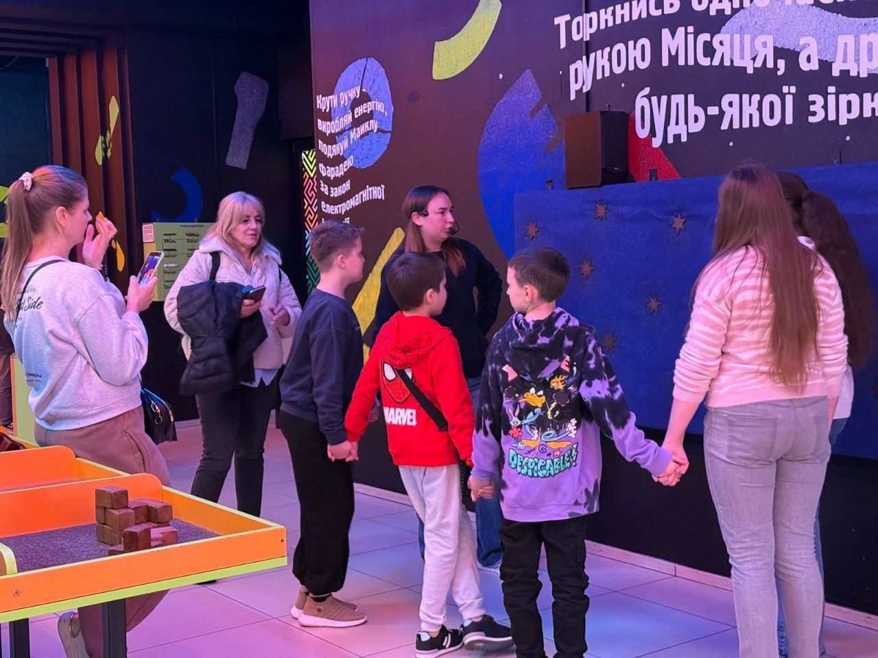 Група дітей та дорослих у науковому центрі біля інтерактивної стіни з написом про космос. Діти тримаються за руки, беручи участь у колективному досліді, а поруч на столі розміщено дерев'яні блоки для конструювання.