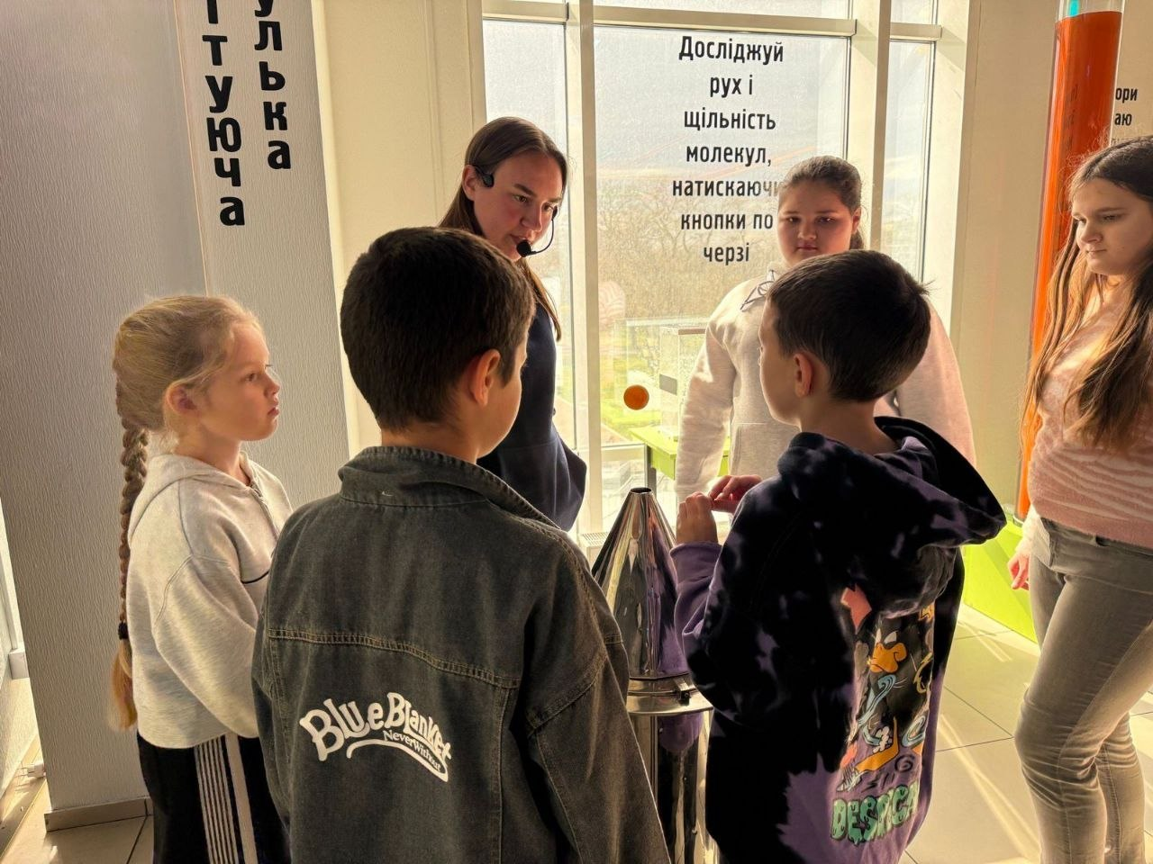 Група дітей слухає пояснення екскурсовода біля інтерактивного експоната в музеї науки. На вікні розміщено напис із закликом досліджувати рух і щільність молекул, натискаючи кнопки.