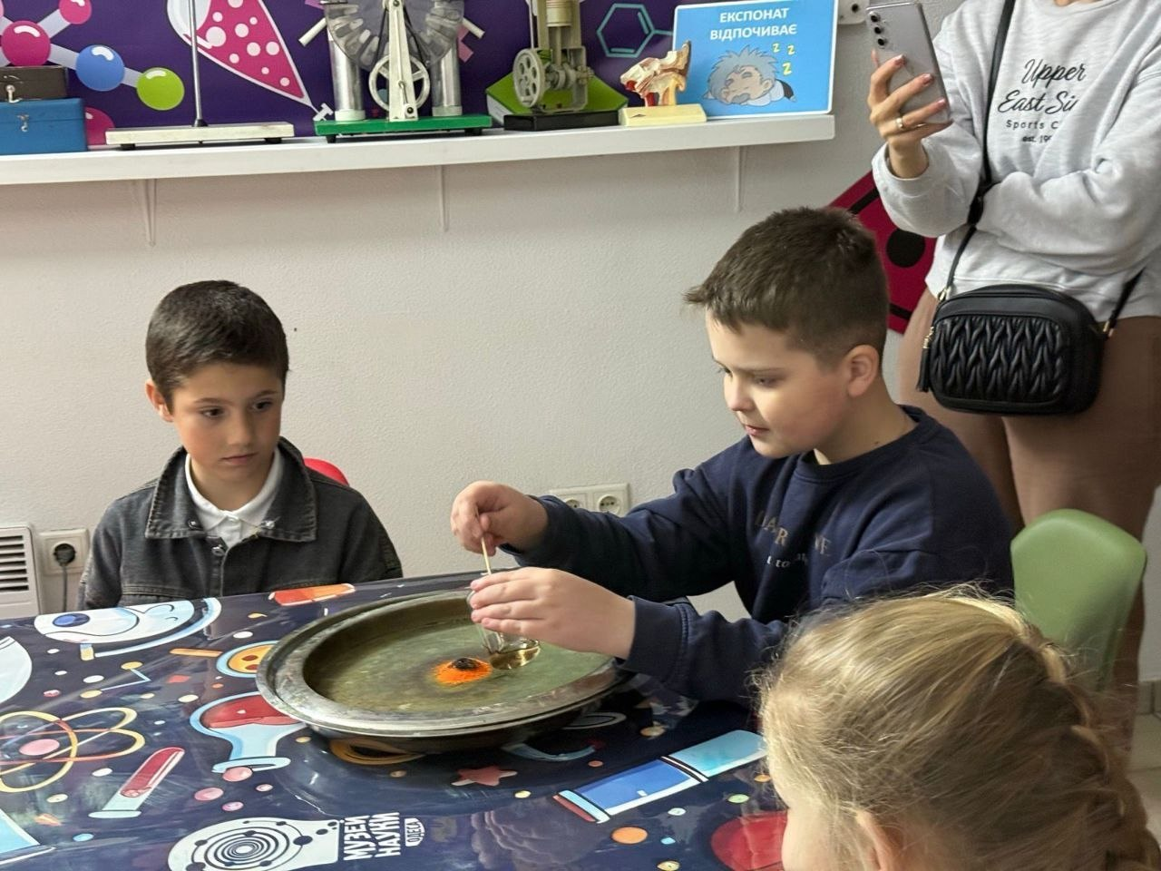 Діти шкільного віку проводять науковий дослід за столом у музеї науки. Хлопчик за допомогою палички взаємодіє з речовиною на металевому таці, а інші діти спостерігають за процесом.