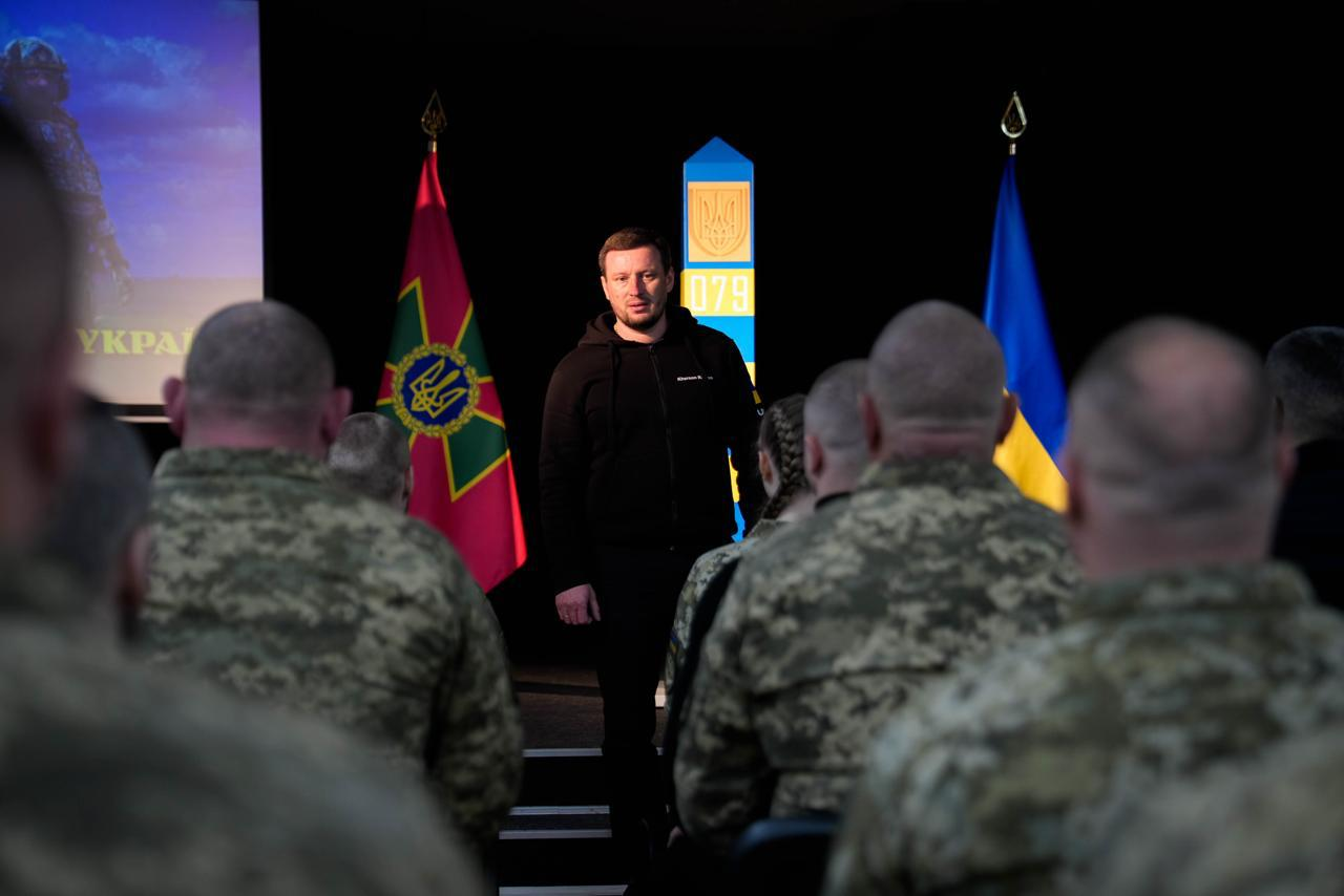 Чоловік у чорному одязі виступає перед групою військовослужбовців у залі. На задньому плані стоять прапори України та Державної прикордонної служби, а також встановлено прикордонний стовп.