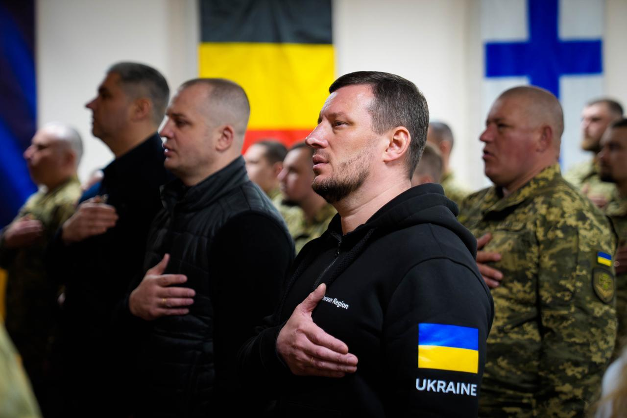 Група чоловіків у цивільному та військовому одязі стоять у шерензі, приклавши праву руку до серця. На фоні видно прапори різних держав, зокрема Бельгії та Фінляндії.