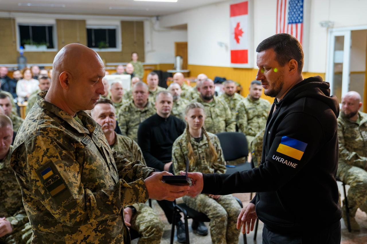 Чоловік у чорному худі вручає нагороду військовослужбовцю — старшому чоловіку міцної статури з поголеною головою, одягненому у піксельну форму з українським шевроном. Церемонія проходить у залі в присутності військових; на стіні позаду висять прапори Канади та США.