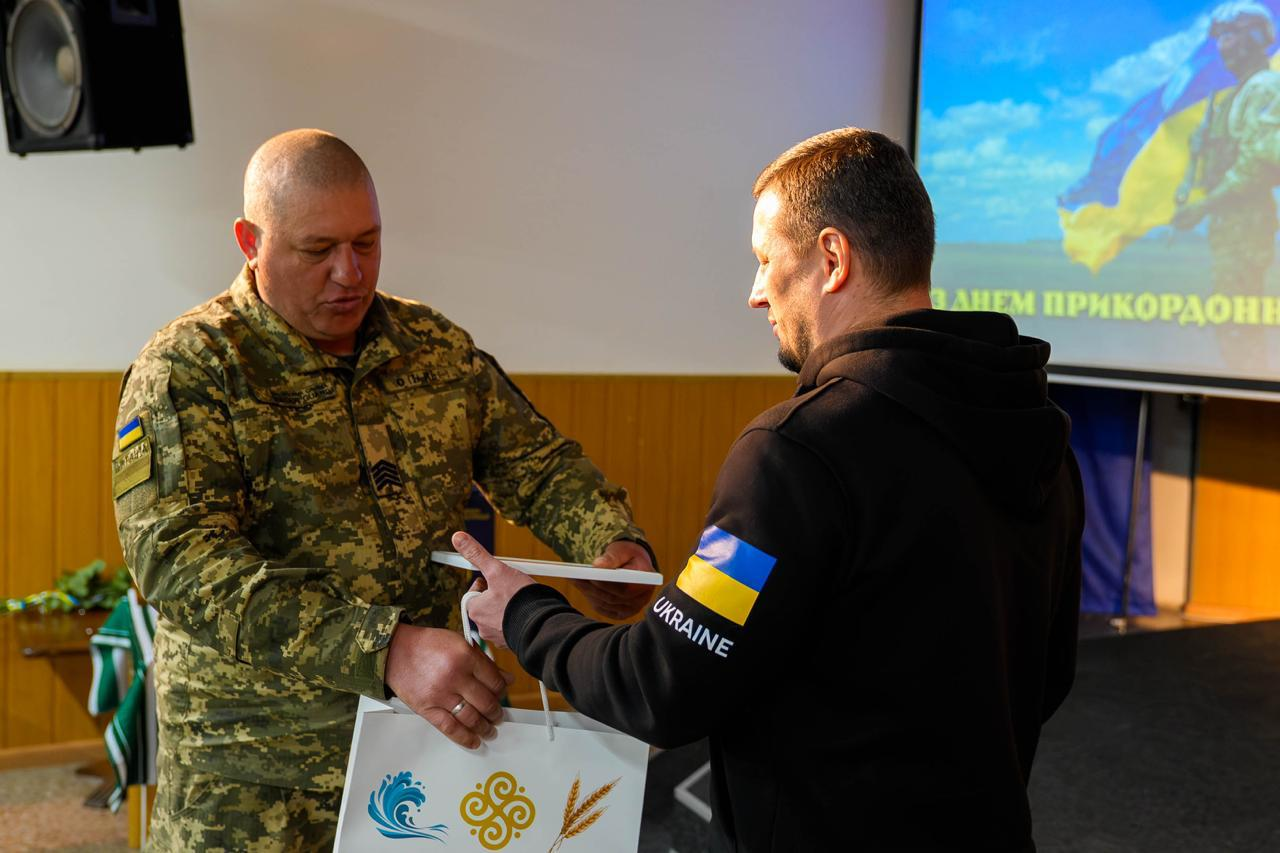 Чоловік у чорному худі вручає грамоту та пакунок військовослужбовцю — чоловіку міцної статури з коротко стриженим волоссям і сивиною на скронях, одягненому у піксельну форму з нашивками. На задньому плані видно екран із привітанням до Дня прикордонника.