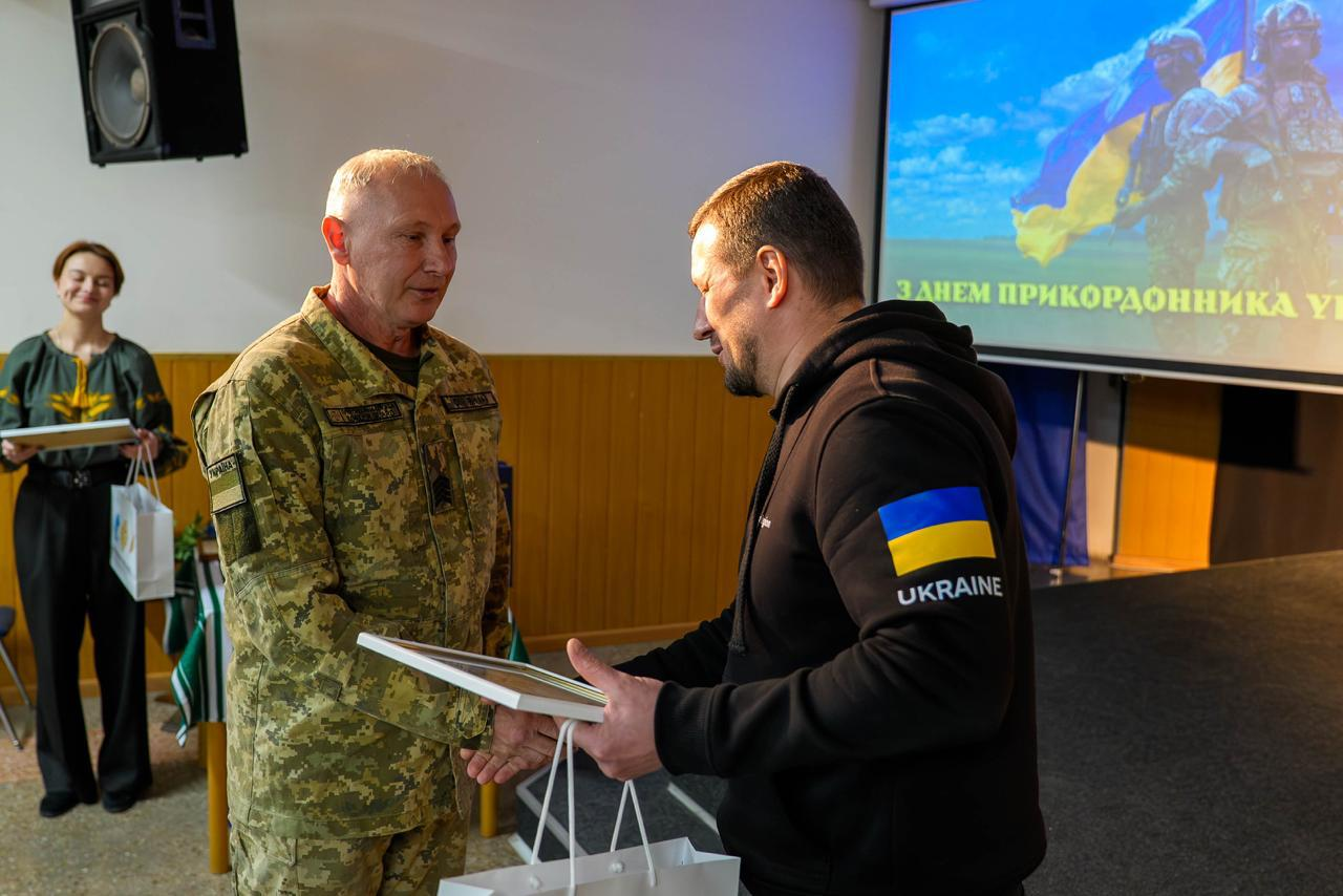 Чоловік у худі вручає грамоту сивочолому військовому на фоні екрана з привітанням до Дня прикордонника. Поруч стоїть дівчина у вишиванці з іншими нагородами в руках.