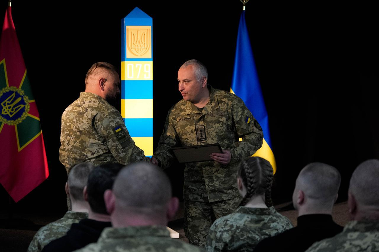 ійськовослужбовець із короткою сивою стрижкою вручає грамоту побратиму — чоловіку з бородою та характерною зачіскою з виголеними скронями. Нагородження відбувається на фоні прикордонного стовпа та прапорів; на передньому плані видно потилиці інших присутніх у залі.