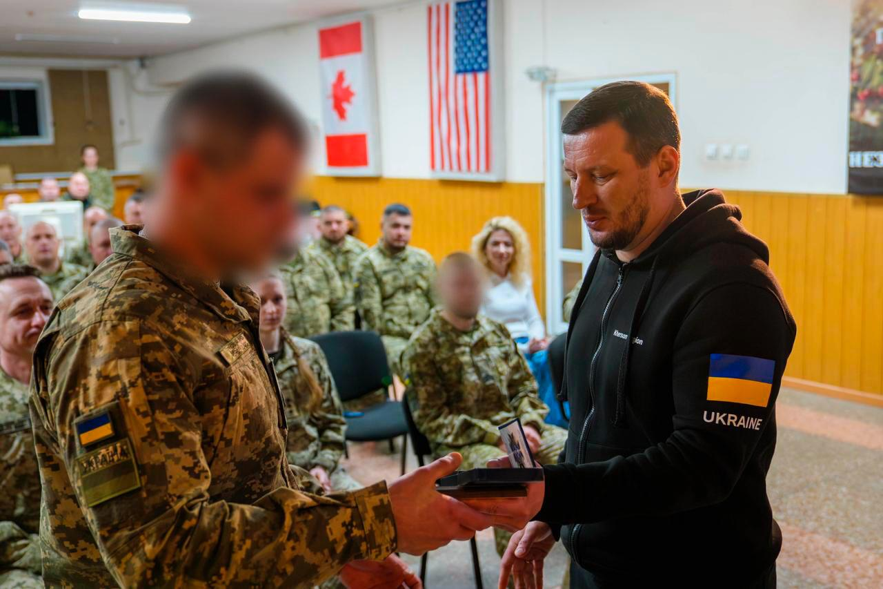 Чоловік у чорному худі вручає нагороду військовослужбовцю середньої статури з короткою стрижкою темного волосся, чиє обличчя на фото розмите. Військовий одягнений у піксельну форму; церемонія проходить у залі на фоні іноземних прапорів та інших присутніх.