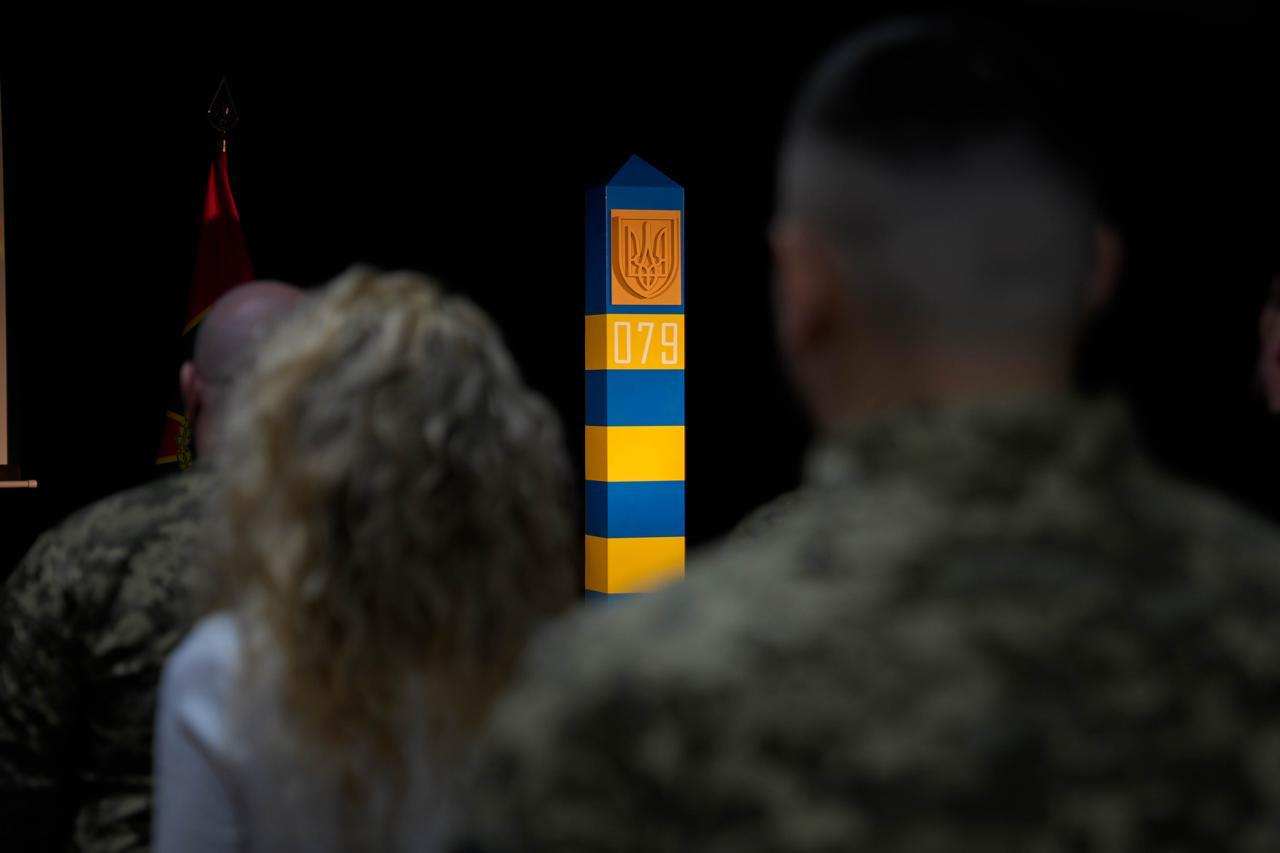 Вигляд крізь плечі присутніх у залі на прикордонний стовп синьо-жовтого кольору з гербом України та номером 079. На передньому плані — потилиці людей у цивільному одязі та військовій формі.