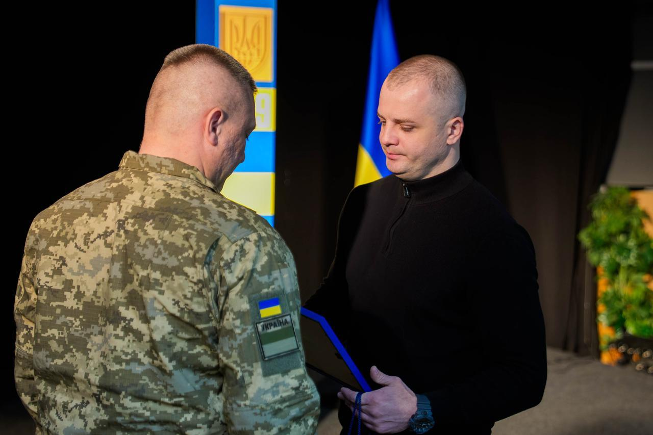 Чоловік середньої статури з короткою стрижкою світлого волосся, одягнений у чорну кофту, тримає синю папку з нагородою. Навпроти нього стоїть військовослужбовець міцної статури з виголеними скронями у піксельній формі (вигляд зі спини). На задньому плані — синьо-жовтий прикордонний стовп.