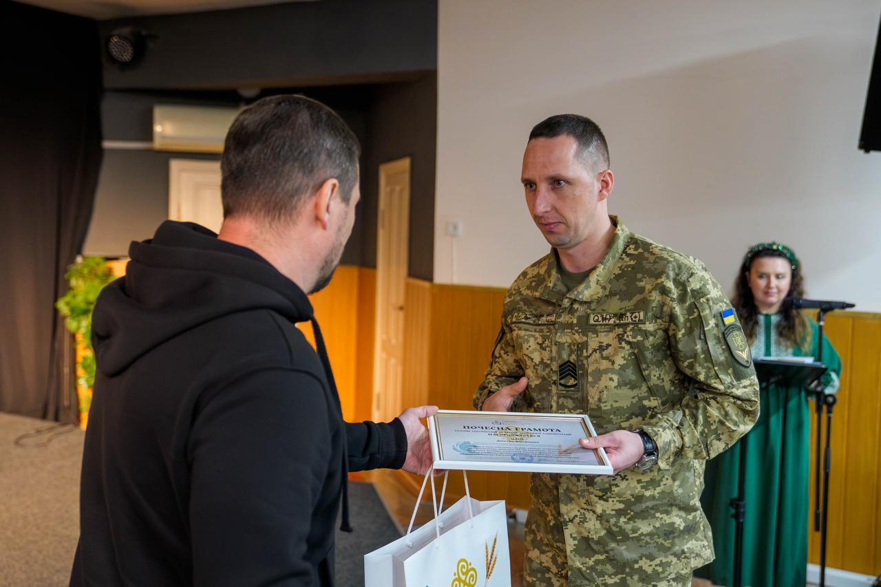 Чоловік у чорному худі вручає «Почесну грамоту» та пакунок військовослужбовцю — високому чоловіку з короткою стрижкою темного волосся та зосередженим виразом обличчя, одягненому у піксельну форму з шевронами. На задньому плані за мікрофоном стоїть дівчина у зеленій сукні та віночку.
