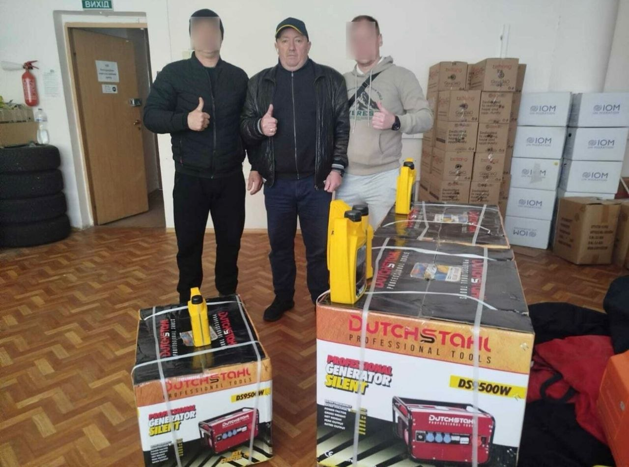 Троє чоловіків стоять у приміщенні біля двох великих коробок із новими генераторами «Dutchstahl». На задньому плані складені численні картонні ящики з логотипом «IOM» (Міжнародна організація з міграції).