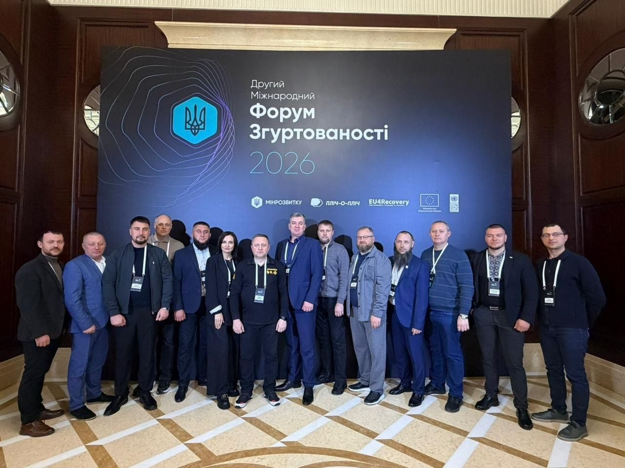 Група чоловіків та одна жінка позують для спільного фото на фоні великого темного банера з написом «Другий Міжнародний Форум Згуртованості 2026». Учасники заходу одягнені в офіційно-діловому та кежуал-стилі, більшість має бейджі на шиї.