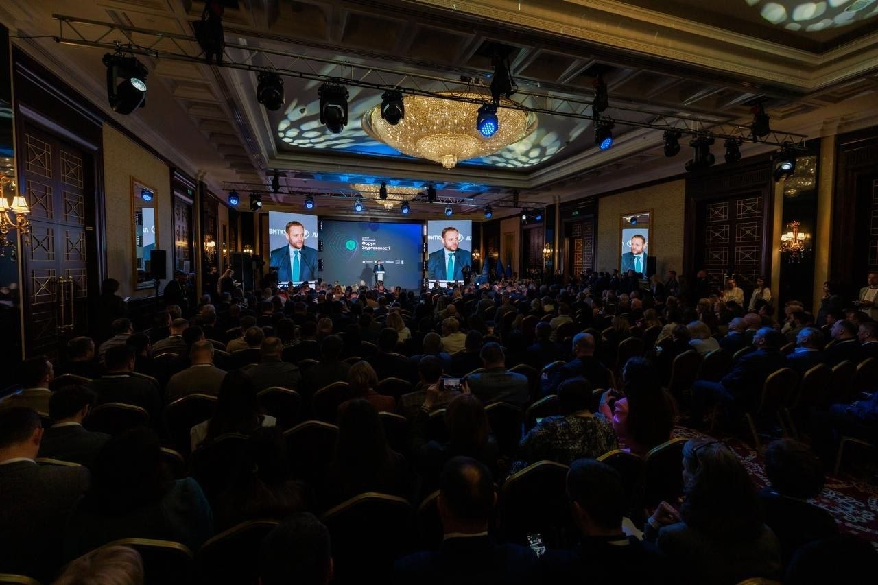 Вигляд із задніх рядів на велику конференц-залу, заповнену людьми під час офіційного форуму. На сцені встановлені великі екрани з трансляцією виступу спікера, а під стелею висить масивна кришталева люстра та професійне освітлювальне обладнання.