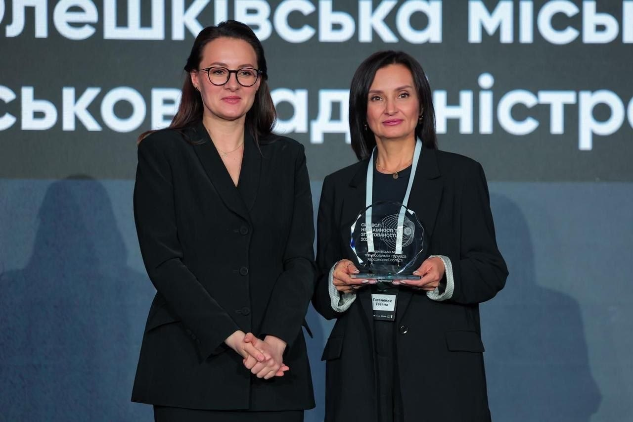 Дві жінки в ділових костюмах позують на фоні банера. Одна з них тримає в руках пам'ятну нагороду зі скла або акрилу. На задньому плані видно частину напису «Олешківська міська військова адміністрація».