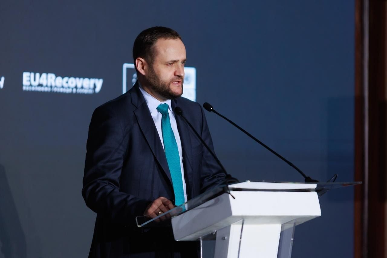 Чоловік у темному костюмі та блакитній краватці виступає за білою трибуною з мікрофонами. На задньому плані — темна стіна з логотипом програми «EU4Recovery».