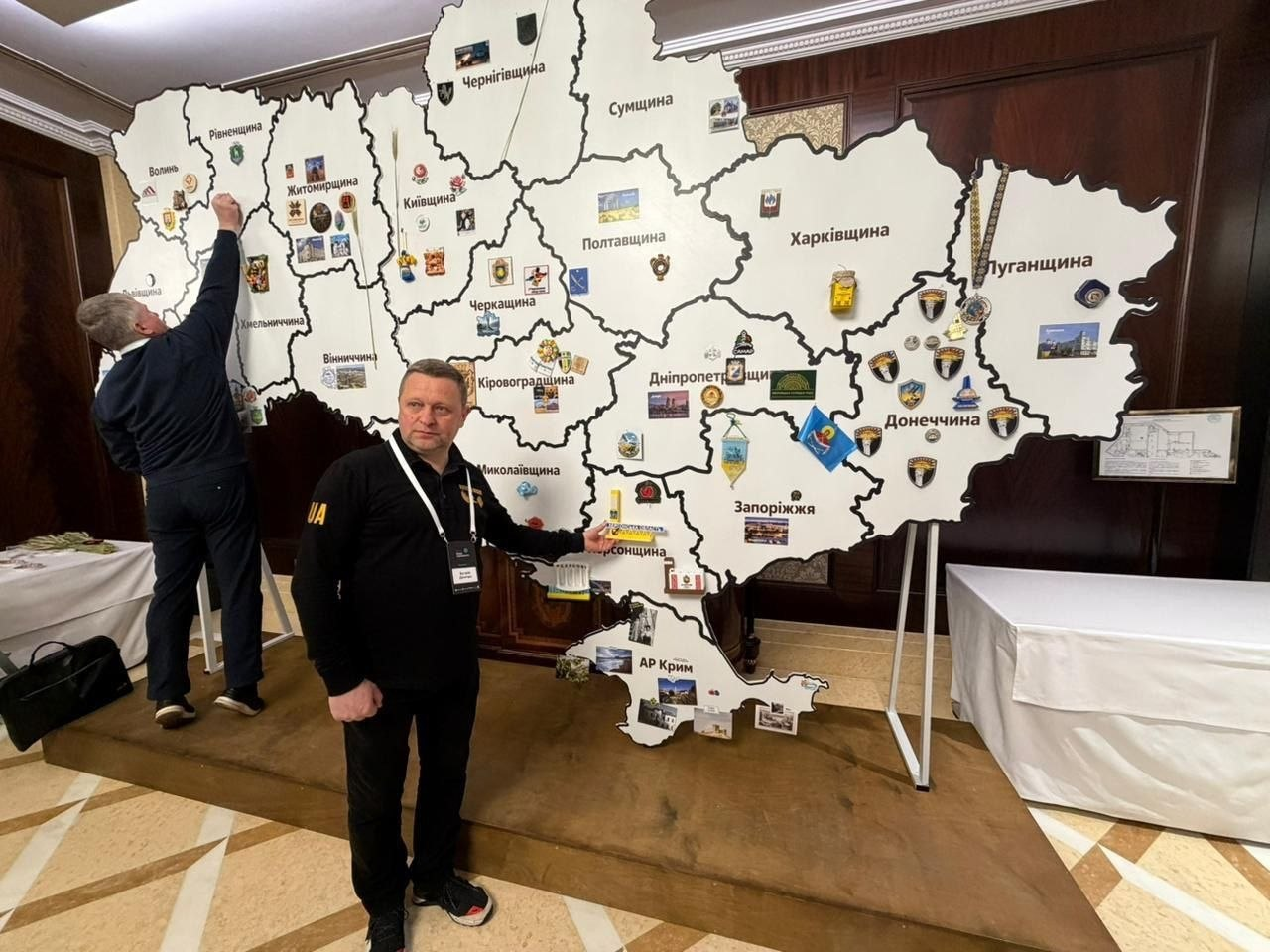 Двоє чоловіків біля великого макета карти України. Один із них стоїть спиною та кріпить позначку на область, а інший вказує рукою на Херсонщину. На макеті зображені межі областей, їхні назви та герби міст.