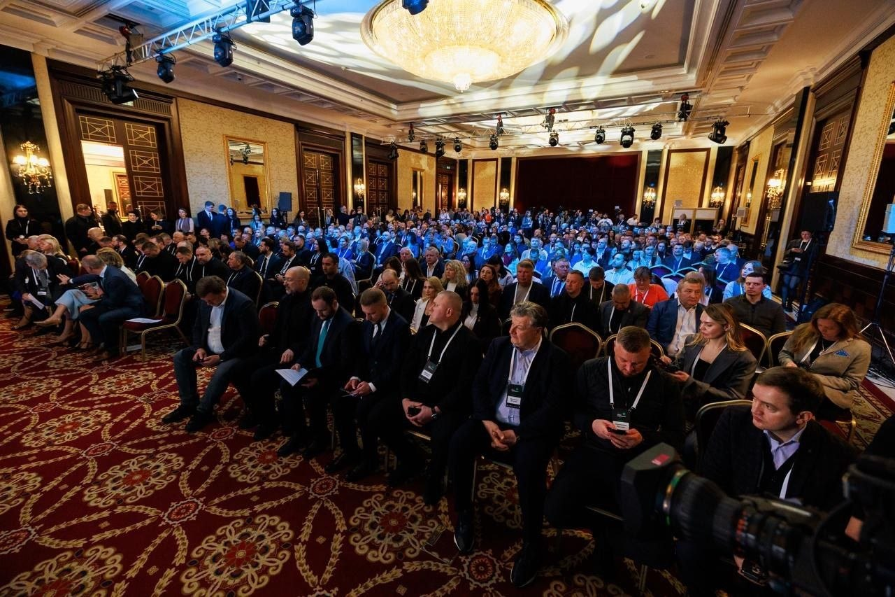 Велика конференц-зала з червоним килимом, заповнена учасниками офіційного заходу. Люди сидять рядами на стільцях, більшість має бейджі; над ними висить масивна кришталева люстра та сценічне освітлення.