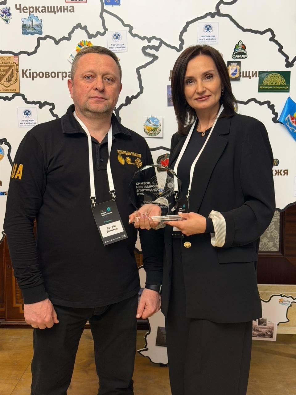 Чоловік у чорному поло та жінка в діловому костюмі разом тримають прозору скляну нагороду на фоні карти України з логотипами міст. На нагороді викарбувано напис «Символ незламності та згуртованості 2025».