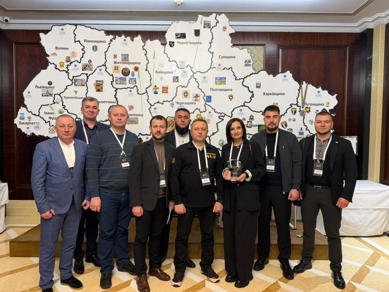 Група чоловіків та одна жінка позують для спільного фото на фоні великої настінної карти України, на якій позначені області та їхні герби. Жінка в центрі тримає в руках прозору скляну нагороду.