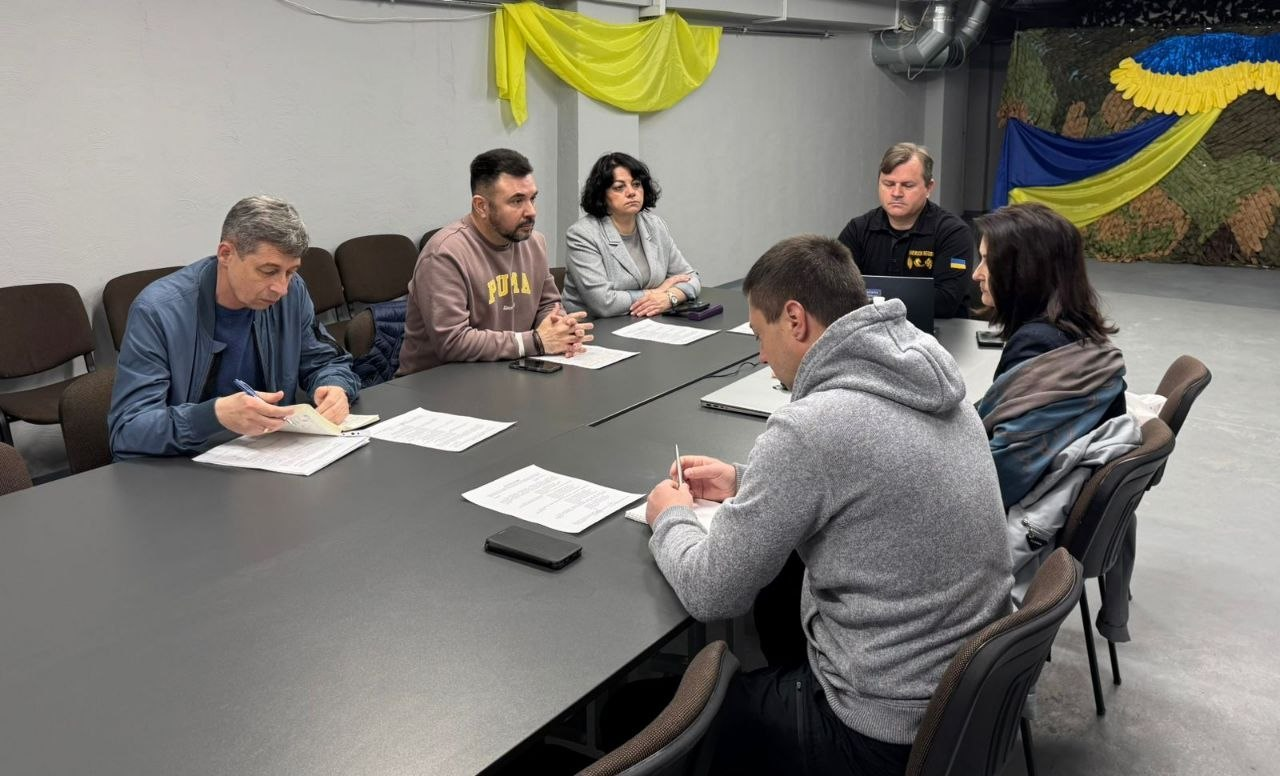 Група людей проводить робочу зустріч за великим столом у приміщенні, схожому на укриття. Учасники переглядають документи та роблять нотатки. На задньому плані стіни прикрашені прапорами України та маскувальною сіткою.