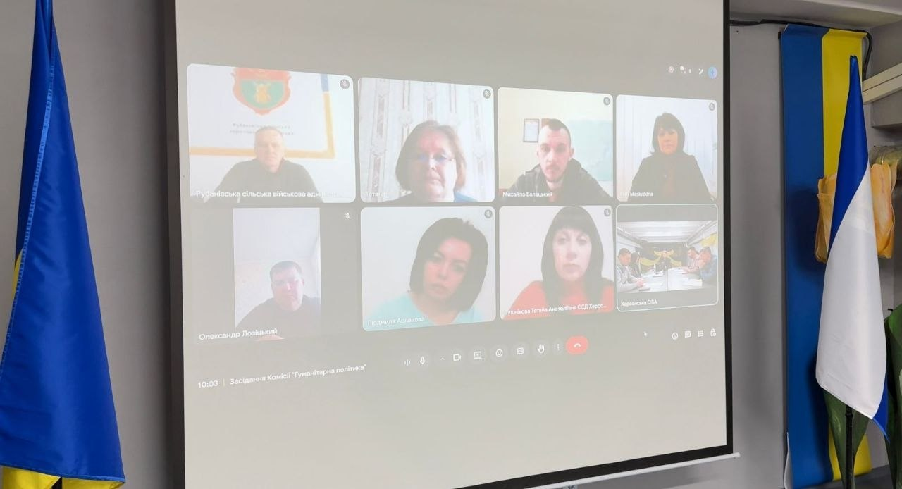 Проєкційний екран у приміщенні, на якому транслюється онлайн-нарада у форматі відеоконференції з вісьмома учасниками. По обидва боки від екрана стоять державний прапор України та прапор міста Херсон.