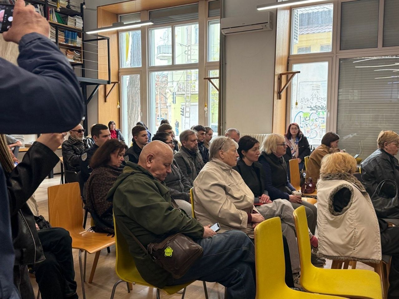 Група людей сидить у залі на фоні великих вікон. Слухачі зосереджені на події, що відбувається перед ними. Ліворуч на передньому плані видно частину постаті людини, яка фотографує на телефон.