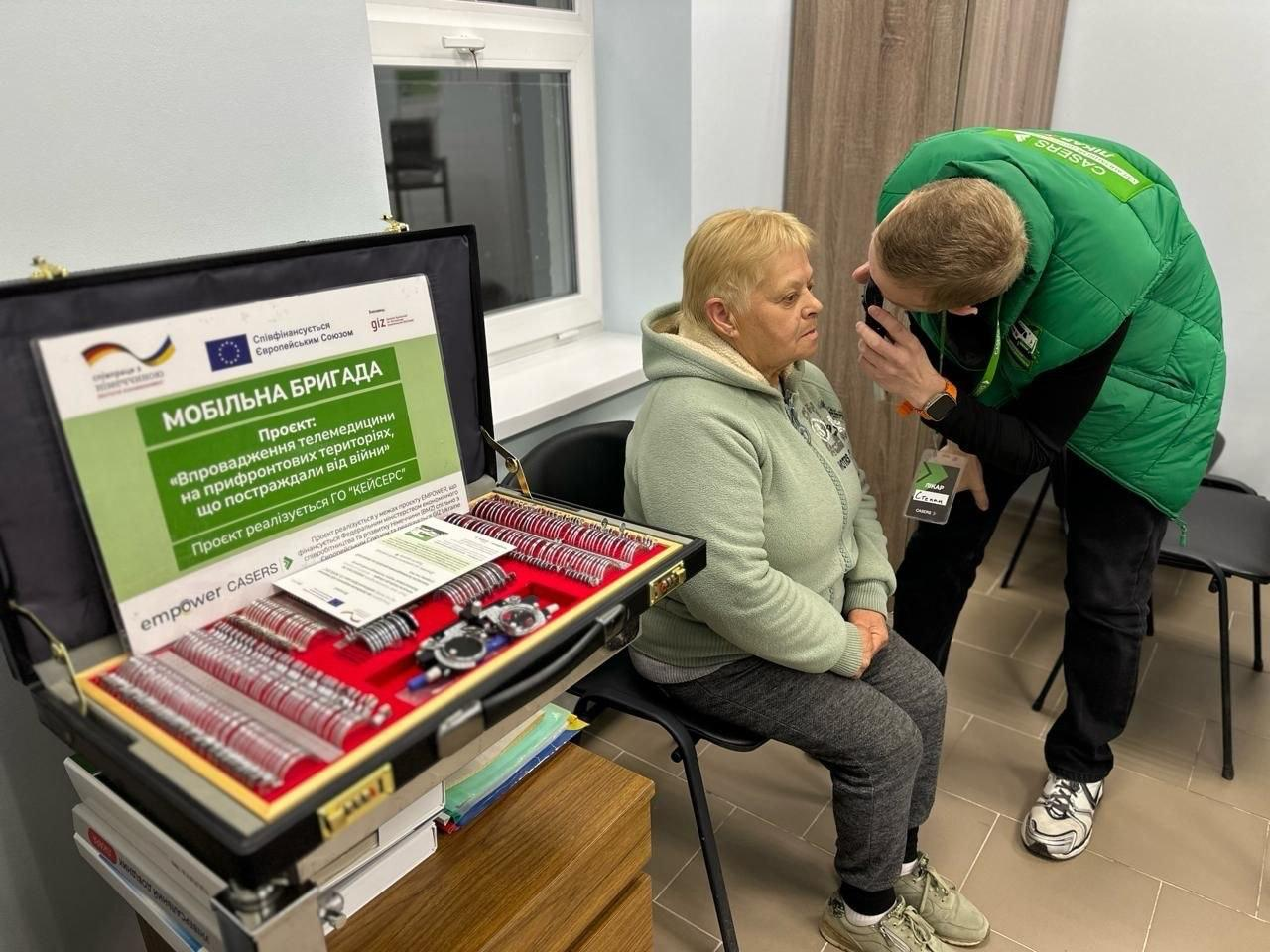 Медичний працівник оглядає очі пацієнтки за допомогою спеціального приладу. Поруч на столі стоїть відкритий кейс із набором лінз для підбору окулярів та інформаційний плакат мобільної бригади.