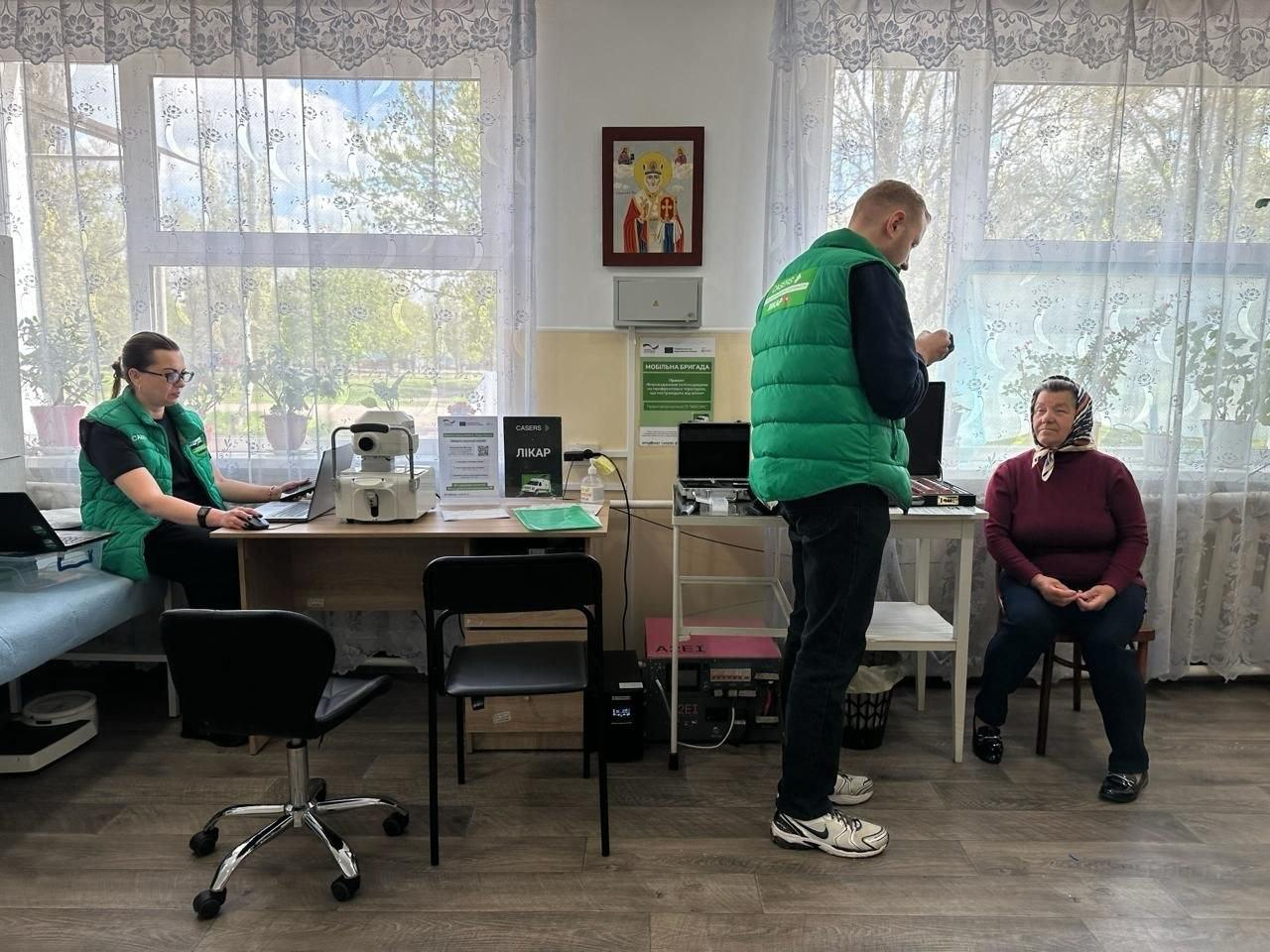 Медичні працівники в зелених жилетах проводять прийом пацієнтки у світлому кабінеті з великими вікнами. На столах розміщене діагностичне обладнання, ноутбуки та медичні кейси. Приміщення оздоблене кімнатними рослинами та іконою на стіні.
