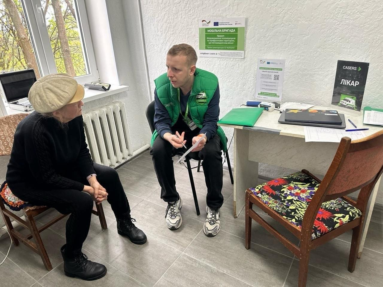 Медичний працівник у зеленому жилеті консультує літню жінку в кабінеті. Вони сидять навпроти один одного біля столу, на якому лежать документи та ноутбук. На стіні розміщено інформаційні плакати мобільної бригади, а поруч видно вікно та радіатор опалення.